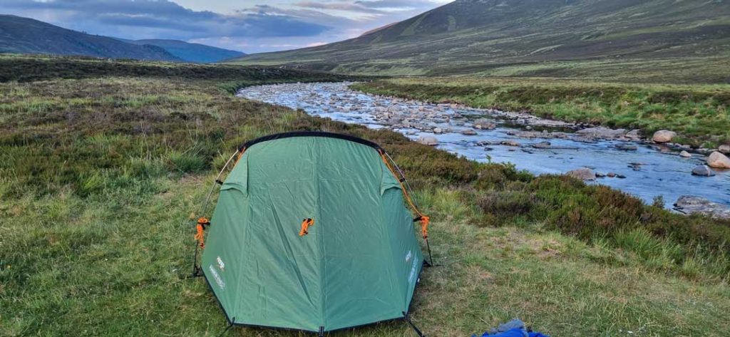 A small two person green tent set up on green grass facing a river that bends around green mountains